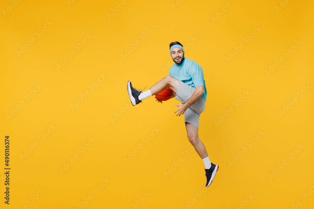 Full body caucasian young fitness trainer instructor sporty man sportsman in headband blue t-shirt hold ball play basketball game jump high isolated on plain yellow background. Workout sport concept.