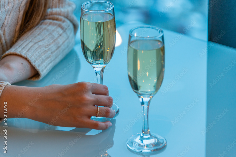 A pair of slender glass goblets with white sparkling wine stands on a glossy table, a woman's hand touches one of the glasses