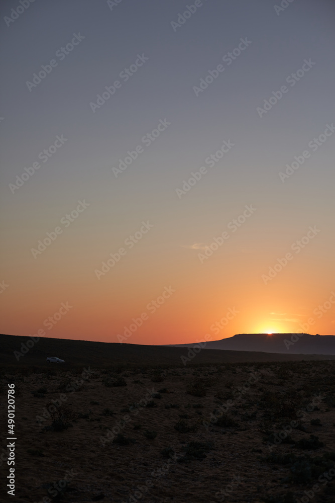 Vertical image of sole white car driving in deserted area catching sunset on its way, sun hiding behind hill leaving orange trace on clear blue sky. Traveling and adventures. Wild nature