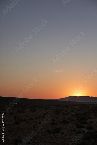 Vertical image of sole white car driving in deserted area catching sunset on its way, sun hiding behind hill leaving orange trace on clear blue sky. Traveling and adventures. Wild nature © Anatoliy Karlyuk