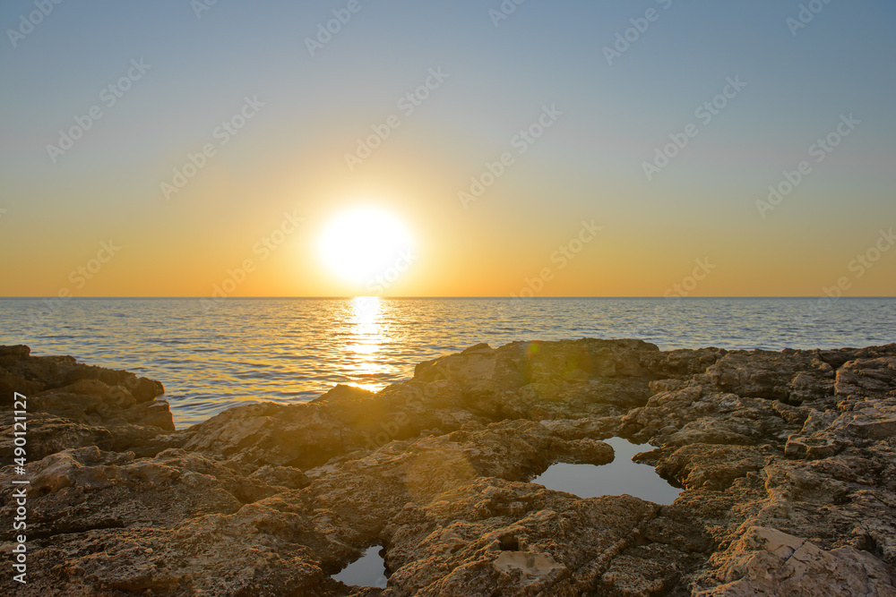 The setting sun on the horizon above the surface of a calm sea with a rocky shore
