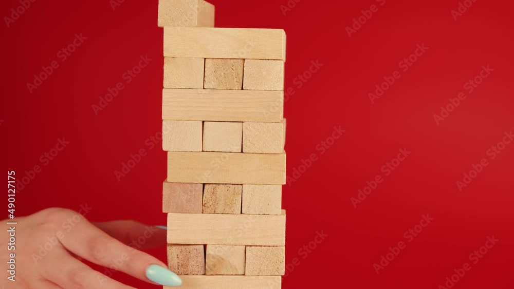 Jenga game on red background. Woman's hand takes out small wooden blocks. Unrecognizable person playing board game Jenga. Concept of leisure activity at home.