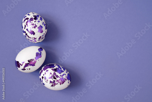Easter eggs decorated with purple foil on a very peri color background with copy space. White painted chicken eggs