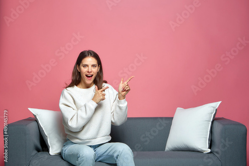 Cool happy young girl point to the side showing on empty space, sitting on couch. Advertising, promotion, sale offer