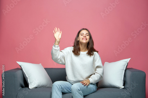 фотографія Smiling beautiful young girl waving hand, greeting with audience or subscribers, sitting on sofa
