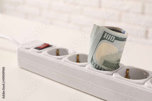 Extension board with rolled dollar banknotes on white table near brick wall, closeup