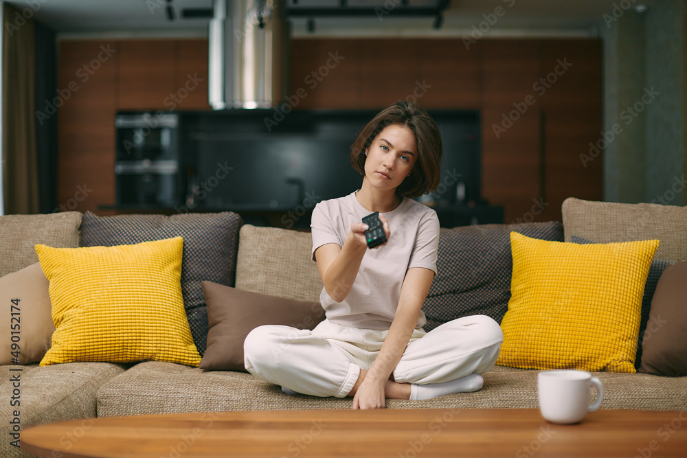 Obraz premium Tired bored young beautiful woman changing tv channels by remote controller, sitting on sofa at home after hard work day