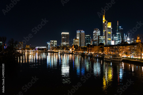 Skyline Frankfurt am Main mit Blick auf den Main von einer Brücke aus. Die Lichter der Büros spiegeln sich auf der Wasseroberfläche. 
