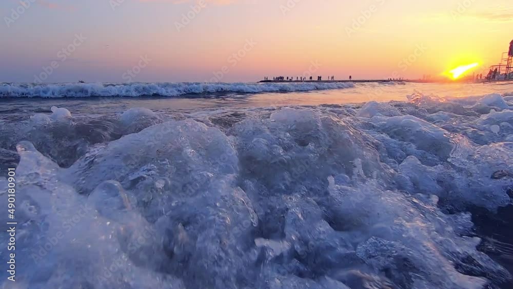 Very low angle shot of sea waves crashing on sandy beach. People walking. Sandy beach, sea coast, sunset dawn sunset sunrise. Sunny path. Seascape, waterscape background. Slow motion. Sea wave inside