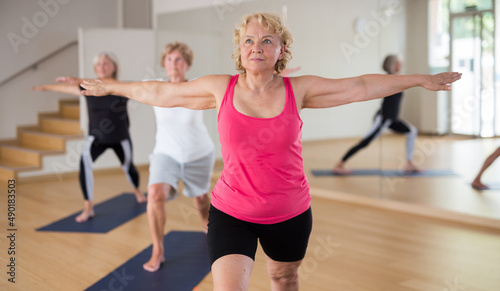 Three mature women doing yoga in a group class perform the exercise by taking the warrior pose II