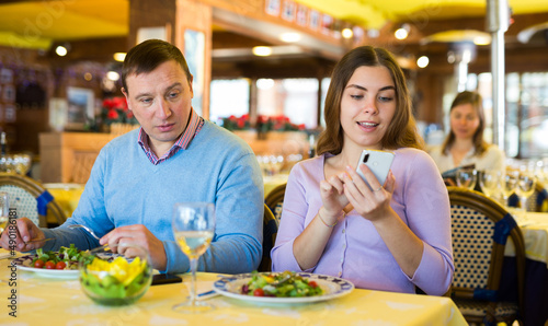 Woman sitting at table with her husband in restaurant and using smartphone.