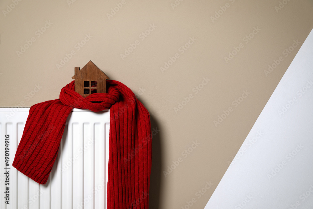 Φωτογραφία Modern radiator with knitted scarf and wooden house near color wall indoors, space for text