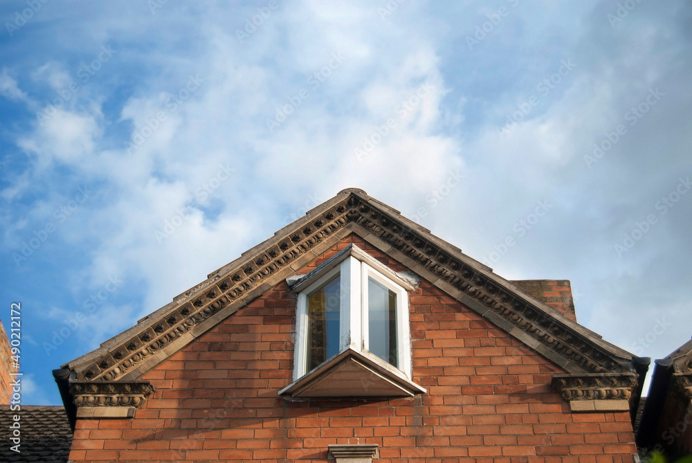 Stone details of a roof peak and a window in a British house view from below against a blue sky with clouds. 