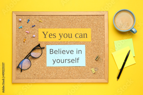 Corkboard with motivational quotes, cup of coffee and office stationery on yellow background, flat lay