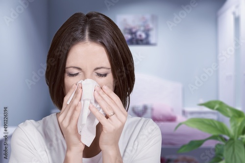 Unhappy sad young female suffering from fever and flu, blowing nose in napkin