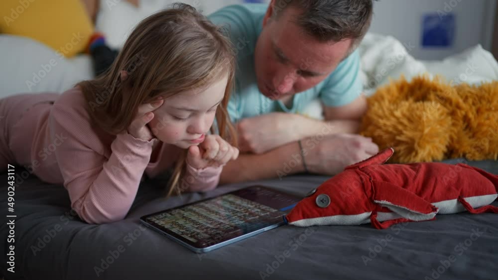 Father and his little daughter with Down syndrome lying on bed and using tablet at home. Father and his little daughter with Down syndrome lying on bed and using tablet at home.