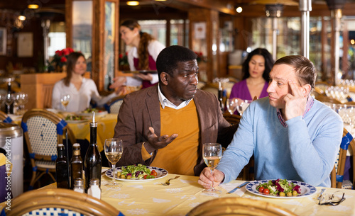 Two friends, African american and Caucasian men, sitting at table in restaurant and having conversation.