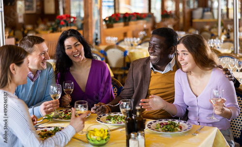 Multiethnic group of positive men and women sitting at table in restaurant  laughing and drinking wine.