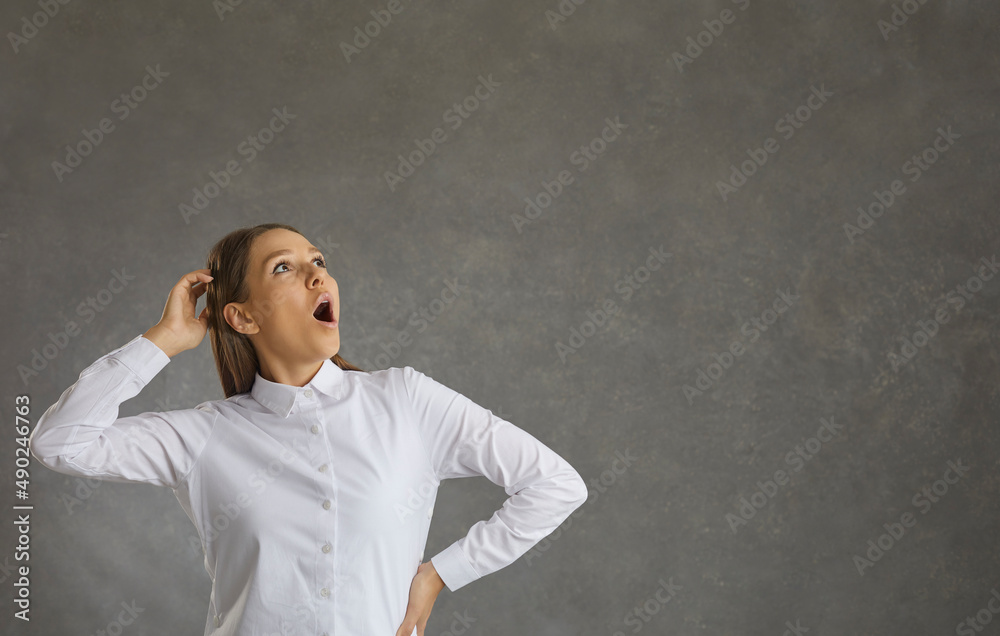 Cheerful woman with a surprised expression looks up while standing on a gray background. Woman with her mouth wide open grabs her head in surprise. Gray background with free space for text. Banner.