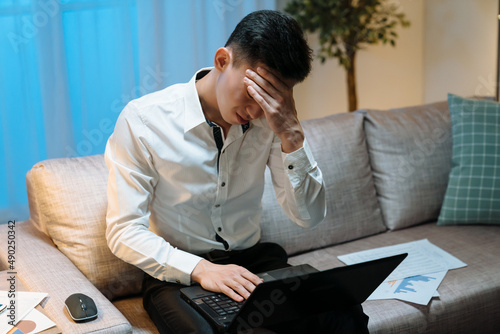 Tired asian japanese man working late in evening in house living room. young guy worker sitting on couch with laptop computer and document charts beside. illness male manager suffer headache in night