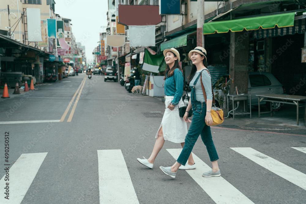 Naklejka premium side view full length of young female people walking around in town. two smiling girl travelers cross zebra road in taipei taiwan. women traveler relax sightseeing in local traditional market outdoor