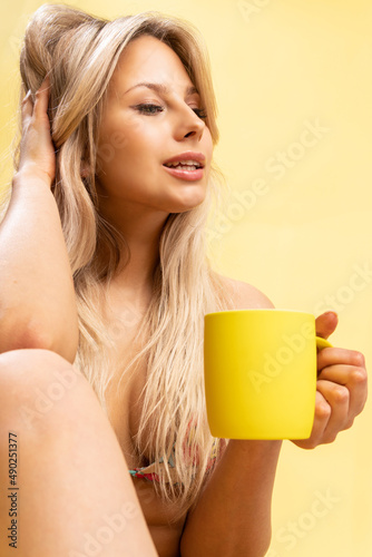 A girl sitting on the floor with her feet cross-legged keeps her head on a beautiful yellow background with a cup in hand yellow or pink and red cheerful beauty  pretty figure slim posing  swimwear