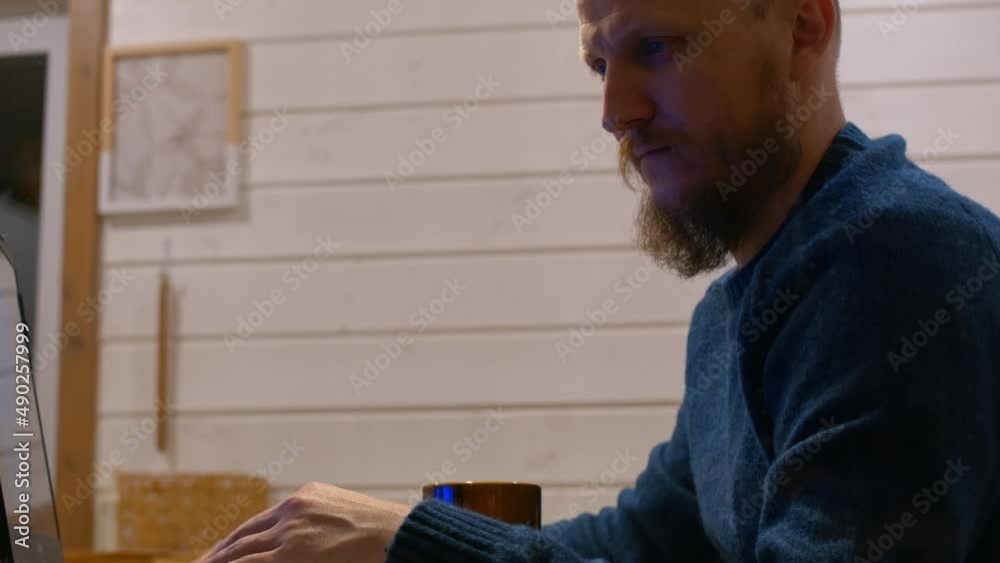 A bearded man sits at a table at home in the evening, works at a computer and drinks coffee from a mug. Bald man at home in the evening view through the window. A bearded man sits at a table at home in the evening, works at a computer and drinks coffee from a mug. Bald man at home in the evening view through the window.