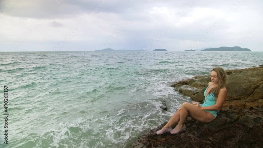 Handsome long-haired woman chill relax resting on rock of sea reef stone, stormy cloudy ocean. Woman in blue swimsuit dress tunic. Concept rest tropical resort coastline traveling outdoor tourism Handsome long-haired woman chill relax resting on rock of sea reef stone, stormy cloudy ocean. Woman in blue swimsuit dress tunic. Concept rest tropical resort coastline traveling outdoor tourism