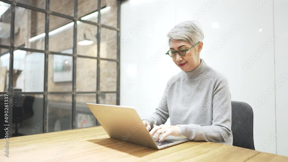Mature asian business woman wears glasses using laptop computer sit at workplace desk. Happy senior older employee 50s businesswoman