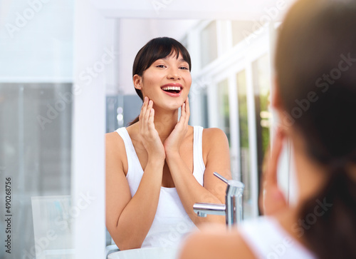 Shes glowing today. Shot of a cheerful attractive young woman applying moisturizer on her face while looking into her reflexion in the mirror.