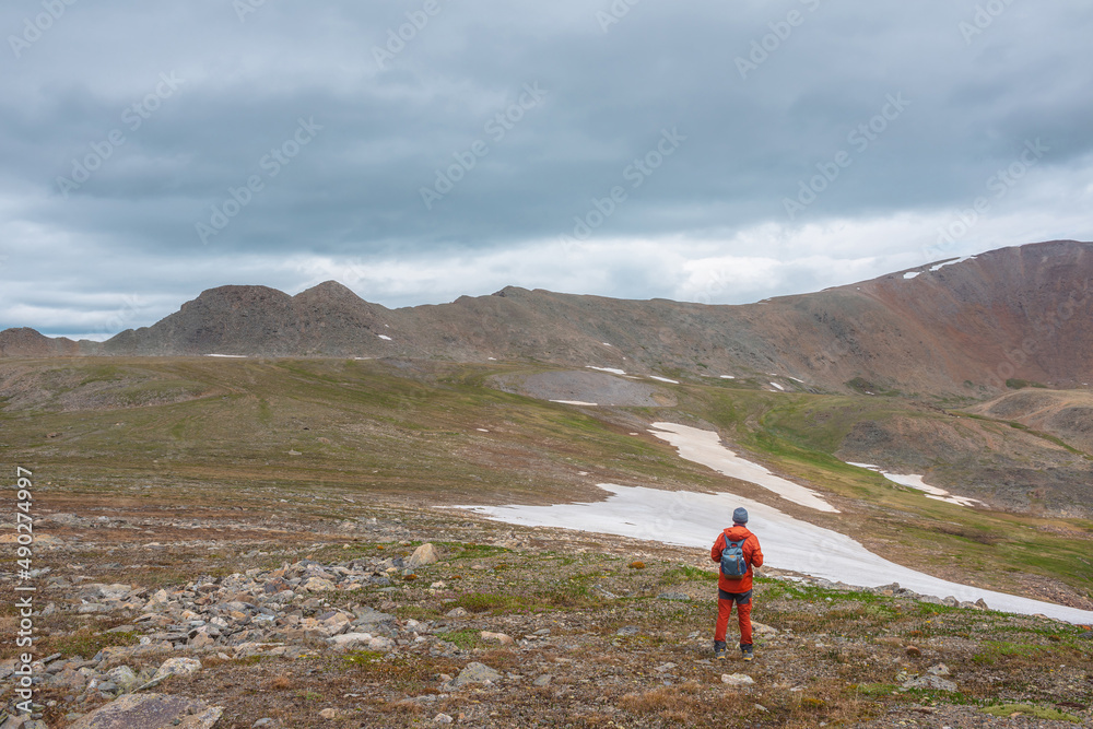 Atmospheric mountain scenery with man in red in grassy stone field with view to large mountain range with sharp rocks under gray cloudy sky. Dramatic mountain landscape with tourist at gloomy weather.