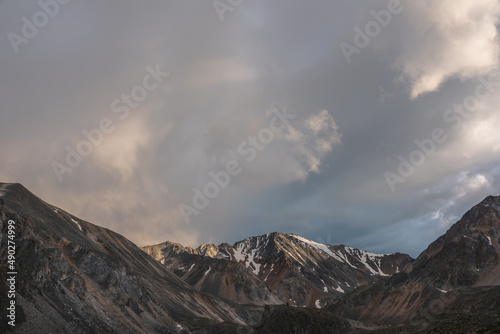 Dramatic landscape with high mountain range with sunlit golden sharp rocky top under clouds of sunset color in gloomy sky. Dark atmospheric scenery with large mountains with snow at changeable weather