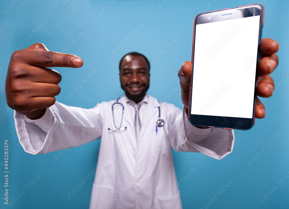 Closeup of smiling medical doctor pointing finger at smartphone with white screen on blue background. Health care professional medic with stethoscope showing mockup on touchscreen device. Closeup of smiling medical doctor pointing finger at smartphone with white screen on blue background. Health care professional medic with stethoscope showing mockup on touchscreen device.