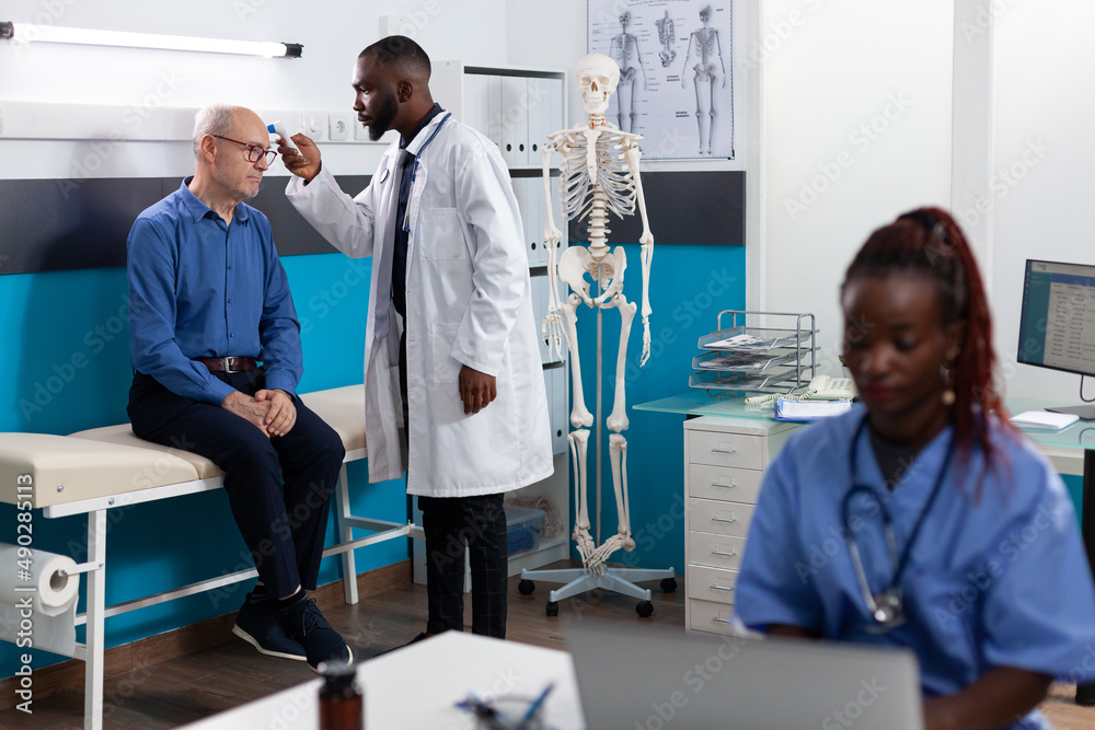 Therapist doctor man measuring temperature using infrared thermometer explaining flu treatment to pensioner man patient during medical consultation in hospital office. Physician scanning forehead Therapist doctor man measuring temperature using infrared thermometer explaining flu treatment to pensioner man patient during medical consultation in hospital office. Physician scanning forehead