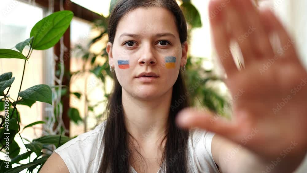 Young Russian-Ukrainian girl with the flag of Ukraine and Russia on her face is saying STOP. The concept of participation of the Ukrainian people in the war with Russia. Not war concept. Young Russian-Ukrainian girl with the flag of Ukraine and Russia on her face is saying STOP. The concept of participation of the Ukrainian people in the war with Russia. Not war concept.
