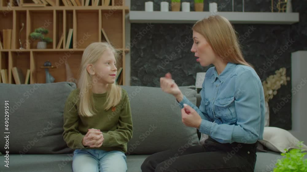 Cute preadolescent daughter with hearing loss and charming hearing impaired mother sitting on couch, friendly chatting with sign language and bonding while happy deaf family enjoying leisure at home.