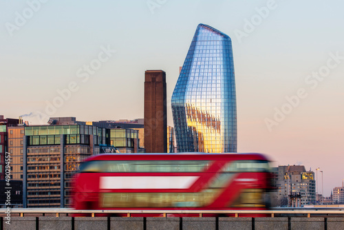 Photography Red London bus driving over London Bridge at sunrise, with tall buildings in the background