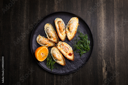 baked mussels with cheese, on a dark plate with herbs and lemon on a wooden background