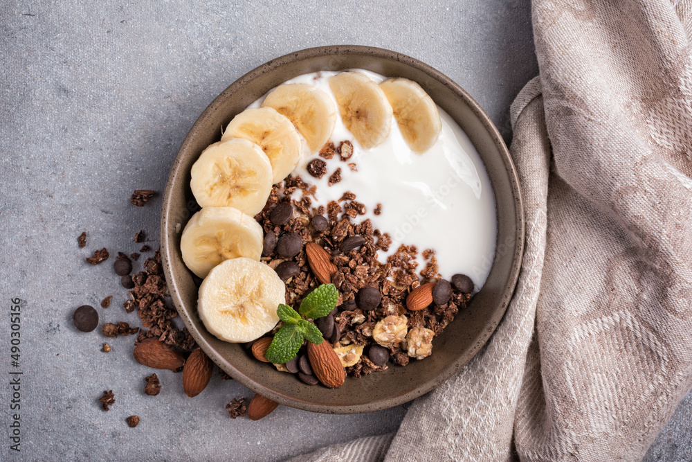 Fototapeta premium Healthy breakfast of granola with fresh banana, nuts, chocolate drops and yogurt