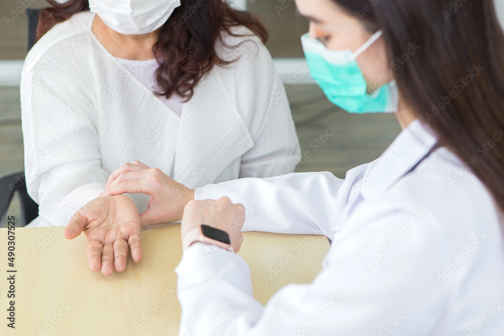 Asian woman doctor takes a pulse of a woman patient while she looks at watch, both wear medical face mask to protect epidemic of COVID-19 (Coronavirus) or pathogen at hospital. Asian woman doctor takes a pulse of a woman patient while she looks at watch, both wear medical face mask to protect epidemic of COVID-19 (Coronavirus) or pathogen at hospital.