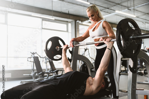 A young male athlete is engaged in a sports club with a woman coach. A man performs a bench press from the chest. The instructor insures the guy.
