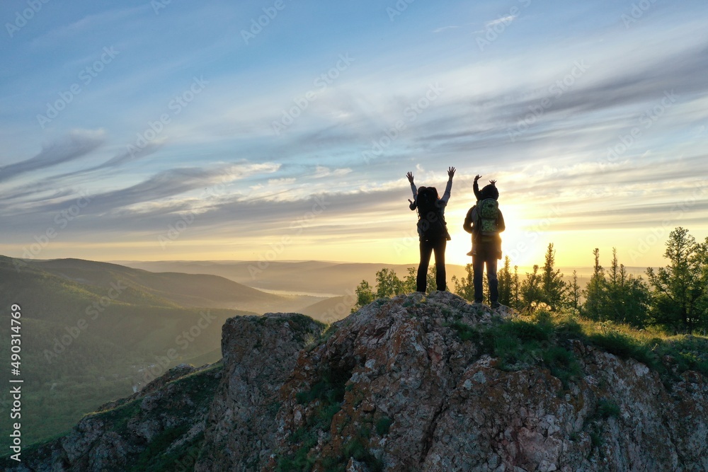 A young couple with two sons happily raise their hands up standing on the top of the mountain at sunset