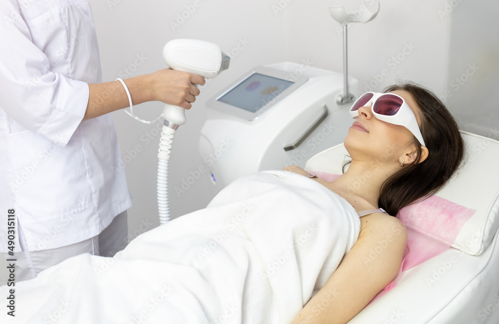 A woman tunes a laser hair removal machine. Girl holding a working part of the epilator in a modern beauty salon. Hair removal, laser procedure at medical clinic, spa concept. Laser skin care banner A woman tunes a laser hair removal machine. Girl holding a working part of the epilator in a modern beauty salon. Hair removal, laser procedure at medical clinic, spa concept. Laser skin care banner