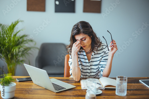 Exhausted businesswoman having a headache in modern office. Mature creative woman working at office desk with spectacles on head feeling tired. Stressed casual business woman feeling eye pain  photo