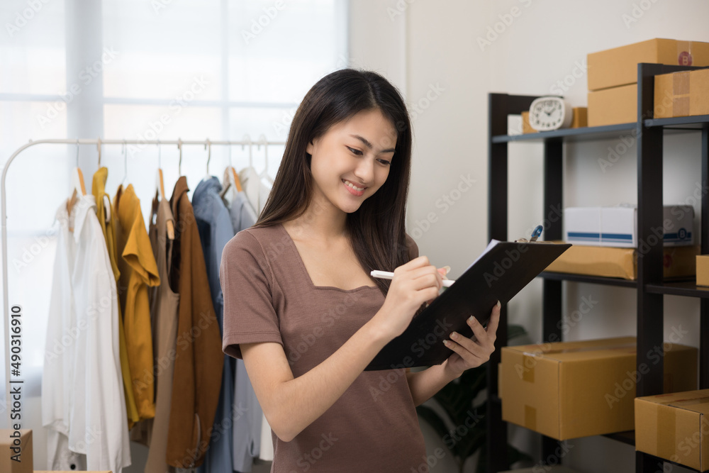 Young beautiful asian woman business owner at fashion store holding clipboard check stock on rails clothing. Asian female entrepreneur working in clothes shop.