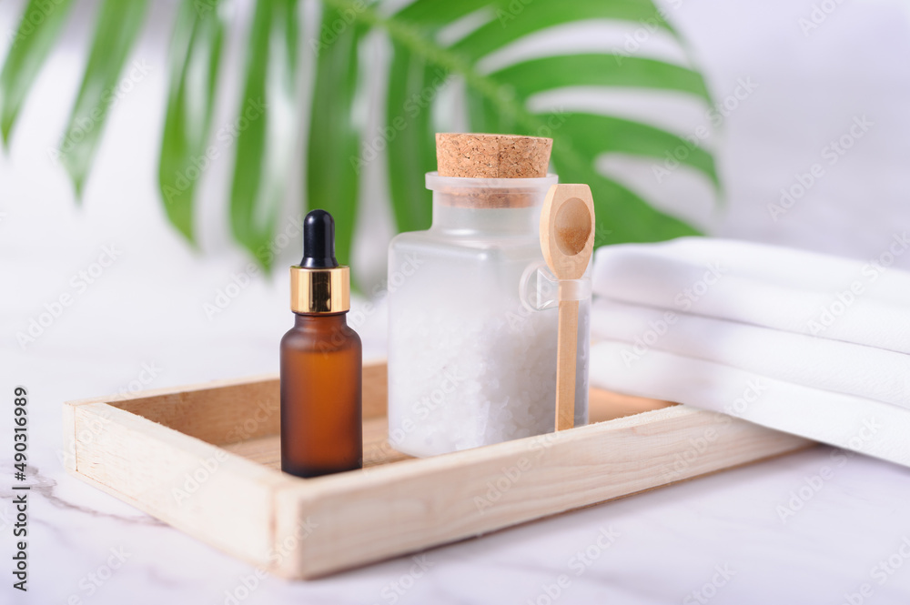 Plastic bottle filled with white bath sea salt and wooden spoon, amber color dropper and white towels on wooden tray.