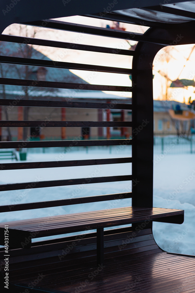 A bench with a wooden dark parallel planks above it. Park during a snowy winter during a sunset