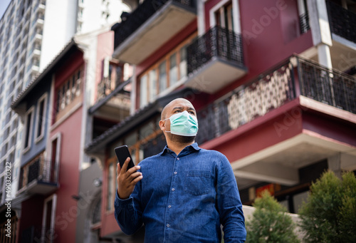 Young indian using smartphone standing in front of office building. Asian man wearing protective mask during pandemic.