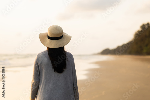 Young asian woman with straw hat standing alone on empty sand beach at sunset seashore. Chilling in holiday weekend summertime. Traveler Female walking around the beach with the sunlight
