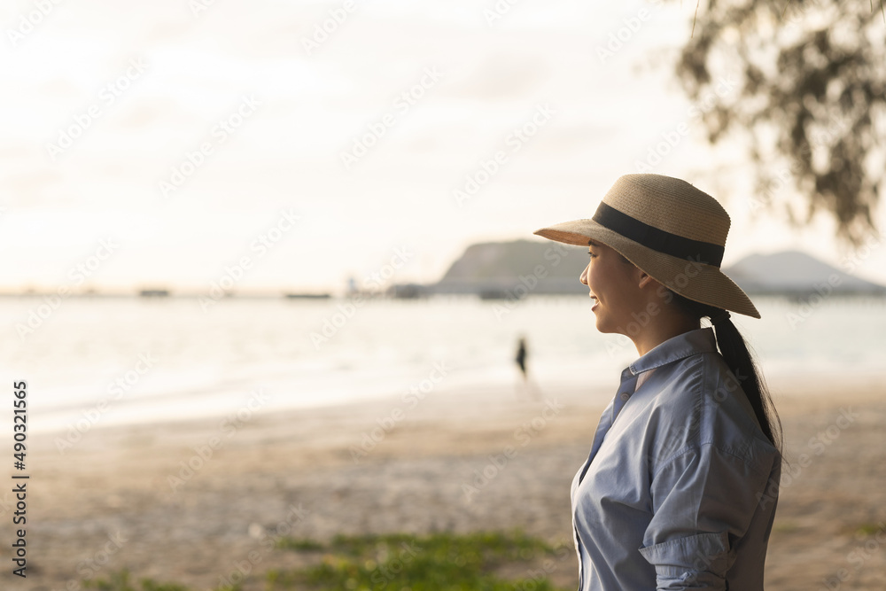 Young asian woman with straw hat standing alone on beach looking at sunset seashore. Chilling in holiday weekend summertime. Traveler Female walking around the beach with the sunlight